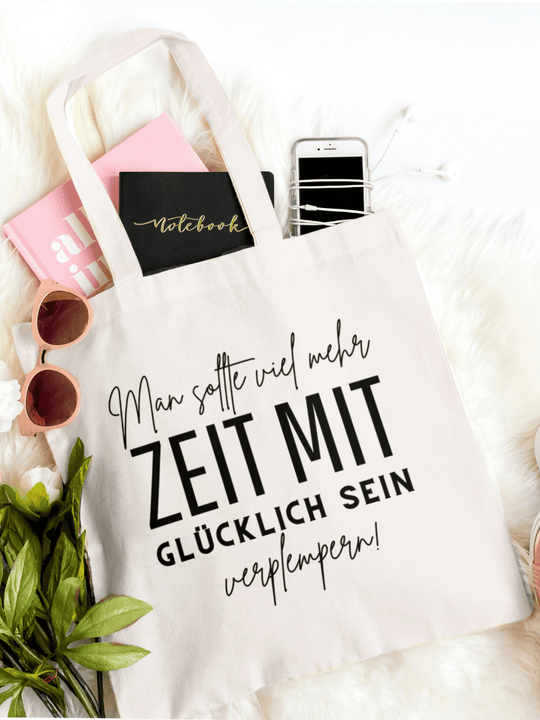 Eine naturfarbene Bio-Baumwolltragetasche, bzw. ein Einkaufsbeutel mit dem Spruch: Man sollte viel mehr Zeit mit glücklich sein verplempern, liegt auf einem Kunstfell mit Blumen, Sonnenbrille, Handy und Buch locker drapiert.
