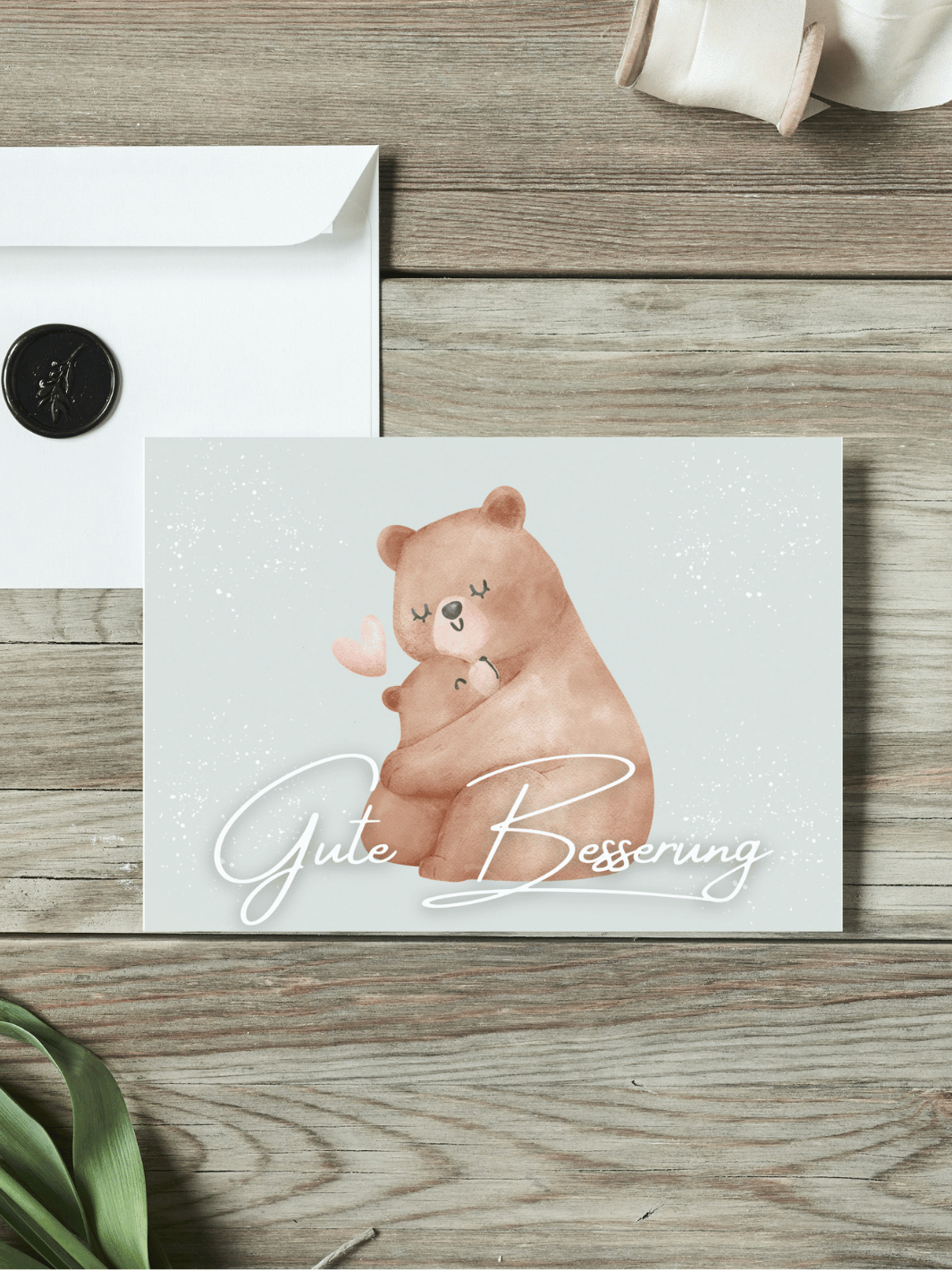 Eine Postkarte mit dem Schriftzug "Gute Besserung" über einem Aquarellmotiv Mutterbär umarmt Babybär liegt auf einem Holztisch. Daneben liegt ein Umschlag mit Wachssiegel, sowie eine alte Schere und Stoffband.