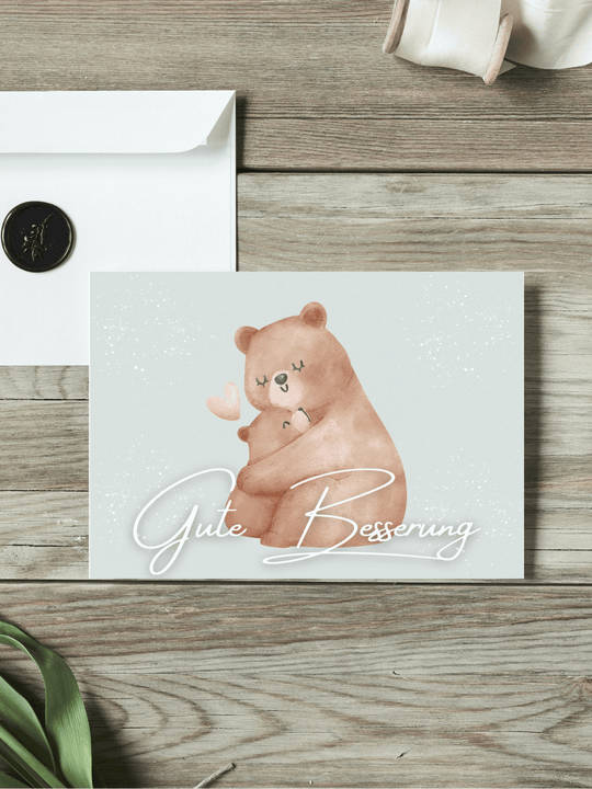 Eine Postkarte mit dem Schriftzug "Gute Besserung" über einem Aquarellmotiv Mutterbär umarmt Babybär liegt auf einem Holztisch. Daneben liegt ein Umschlag mit Wachssiegel, sowie eine alte Schere und Stoffband.