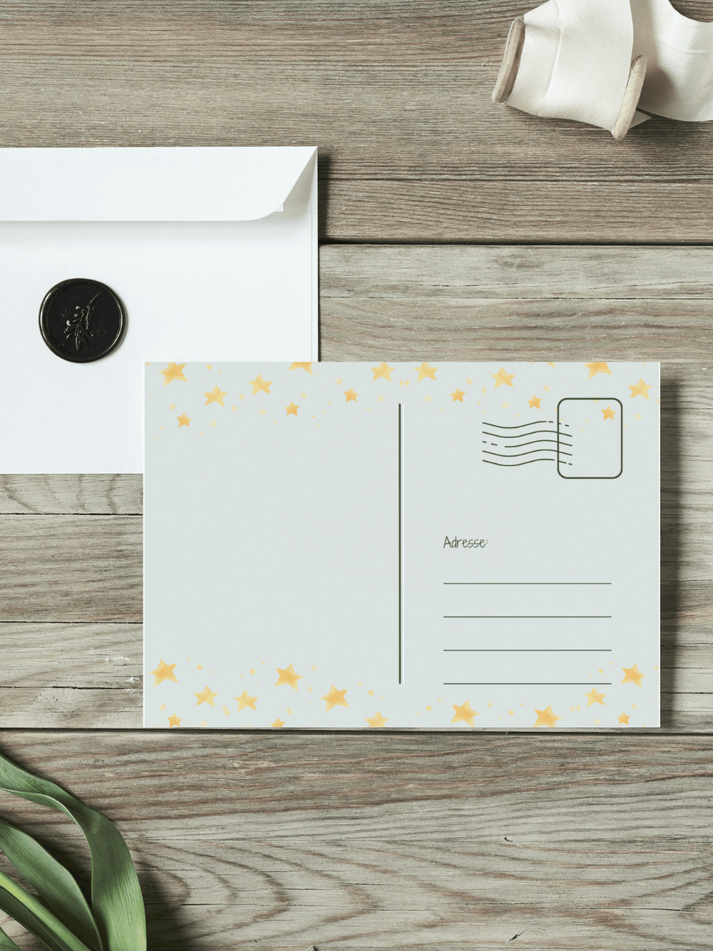 Eine Postkarte mit der Rückseite nach oben liegt auf einem Holztisch. Daneben liegt ein Umschlag mit Wachssiegel, sowie eine alte Schere und Stoffband.