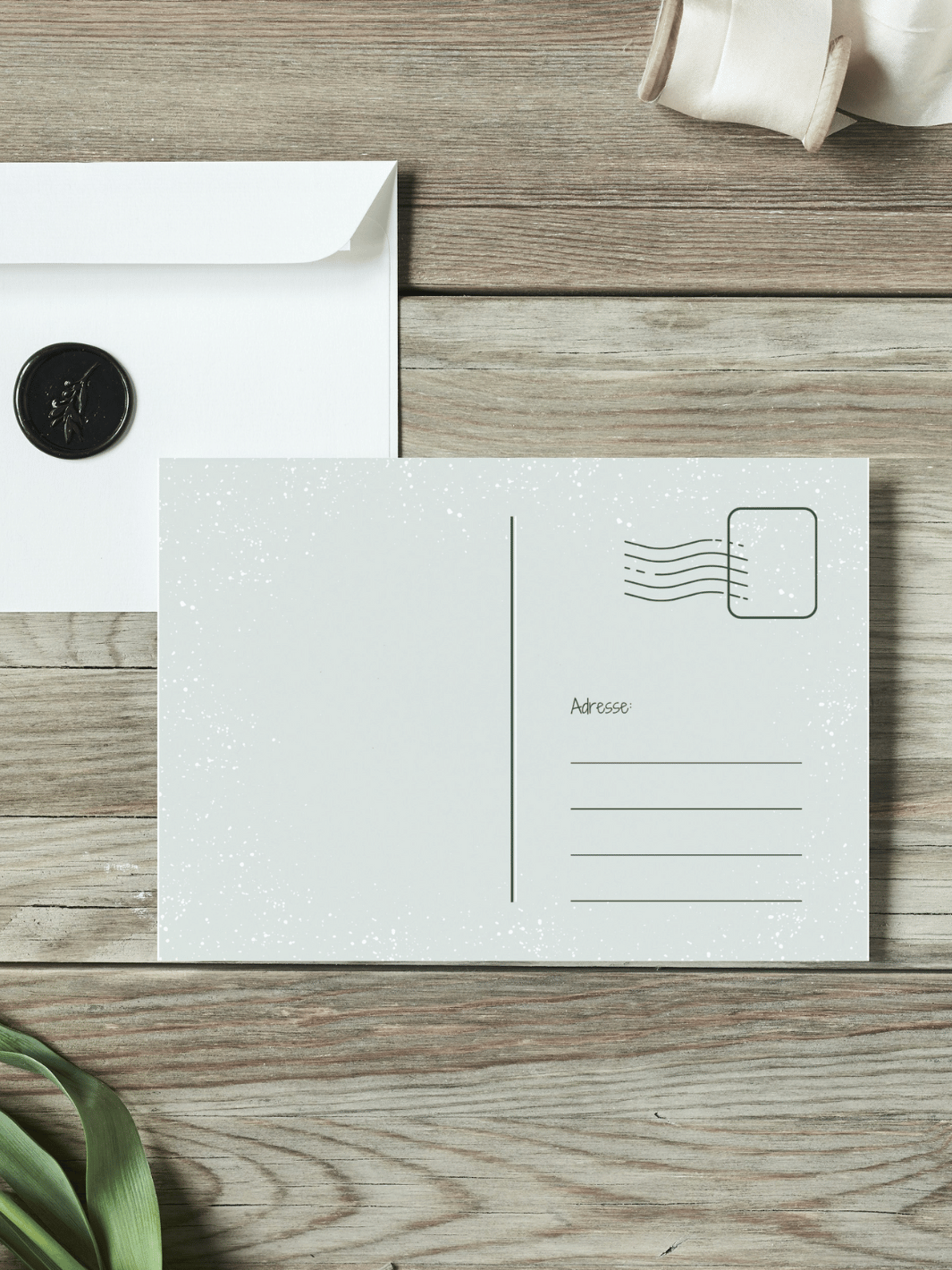 Eine Postkarte mit der Rückseite nach oben liegt auf einem Holztisch. Daneben liegt ein Umschlag mit Wachssiegel, sowie eine alte Schere und Stoffband.