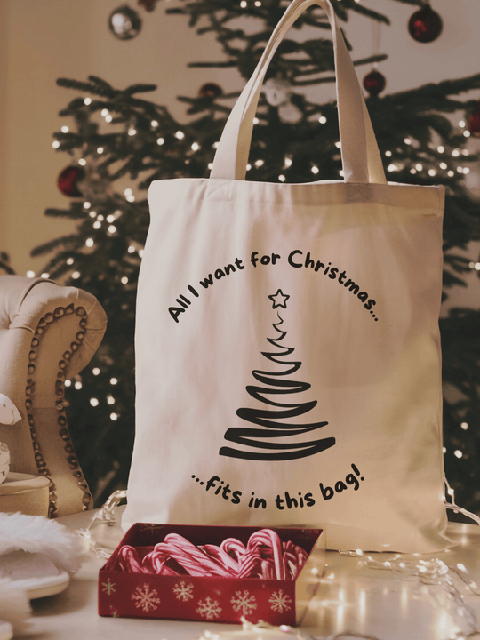 Eine naturfarbene Bio-Baumwolltragetasche, bzw. ein Einkaufsbeutel mit dem Motiv gezeichneter Weihnachtsbaum und dem Spruch: All i want for christmas fits in this bag steht vor einem Weihnachtsbaum mit Zuckerstangen davor