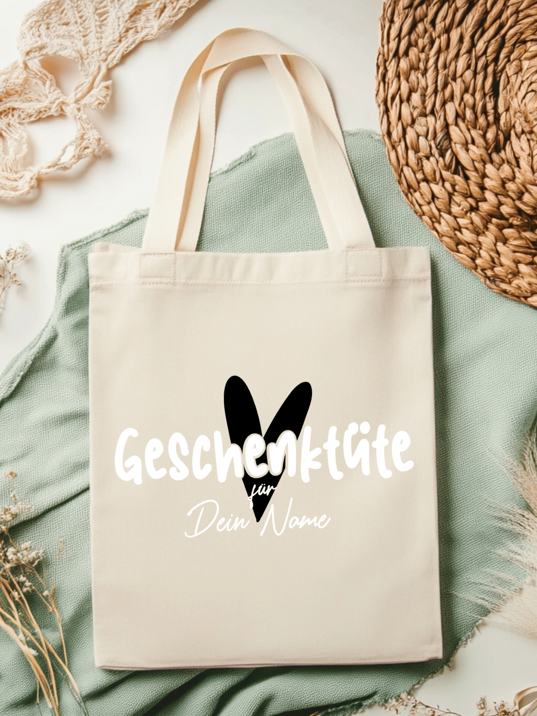 Eine naturfarbene Bio-Baumwolltragetasche, bzw. ein Einkaufsbeutel mit dem Motiv Herz in schwarz und Schriftzug Geschenktüte für Dein Name (personalisierbar) , liegt auf einer salbeifarbenen Stoffbahn locker drapiert.