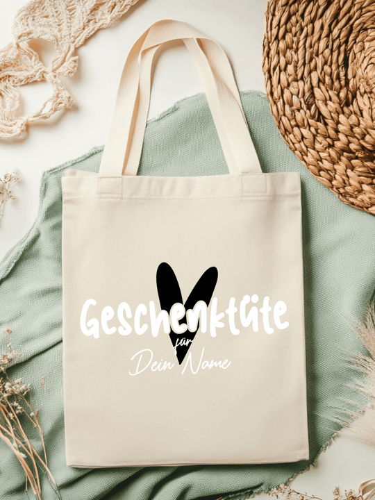 Eine naturfarbene Bio-Baumwolltragetasche, bzw. ein Einkaufsbeutel mit dem Motiv Herz in schwarz und Schriftzug Geschenktüte für Dein Name (personalisierbar) , liegt auf einer salbeifarbenen Stoffbahn locker drapiert.