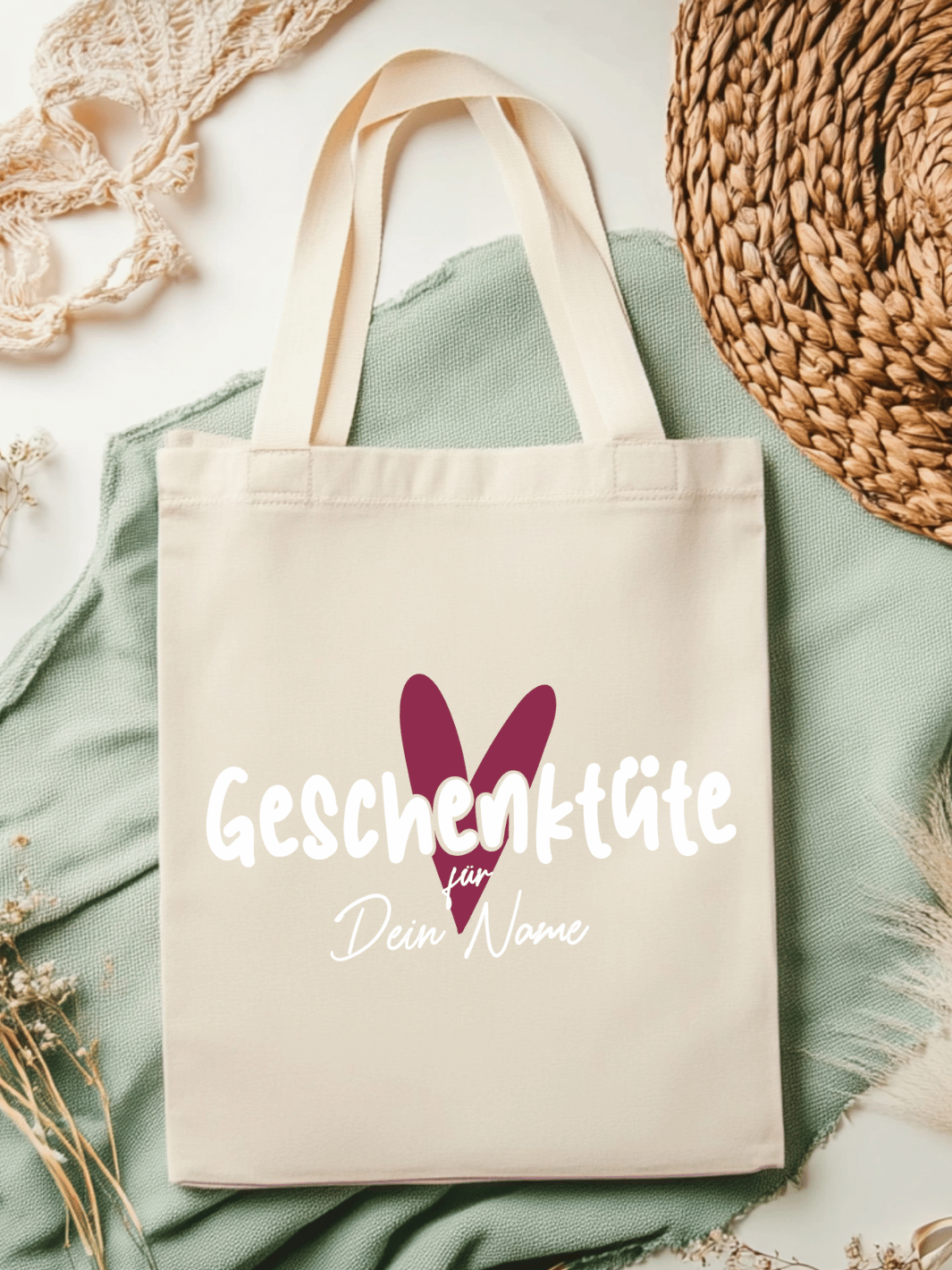 Eine naturfarbene Bio-Baumwolltragetasche, bzw. ein Einkaufsbeutel mit dem Motiv Herz in burgunderrot und Schriftzug Geschenktüte für Dein Name (personalisierbar) , liegt auf einer salbeifarbenen Stoffbahn locker drapiert.