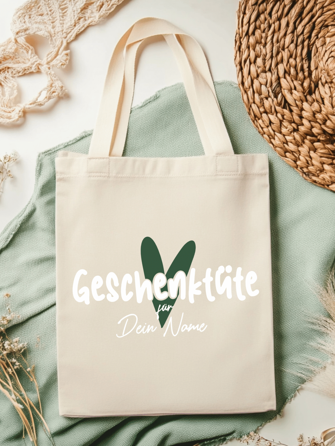 Eine naturfarbene Bio-Baumwolltragetasche, bzw. ein Einkaufsbeutel mit dem Motiv Herz in grün und Schriftzug Geschenktüte für Dein Name (personalisierbar) , liegt auf einer salbeifarbenen Stoffbahn locker drapiert.