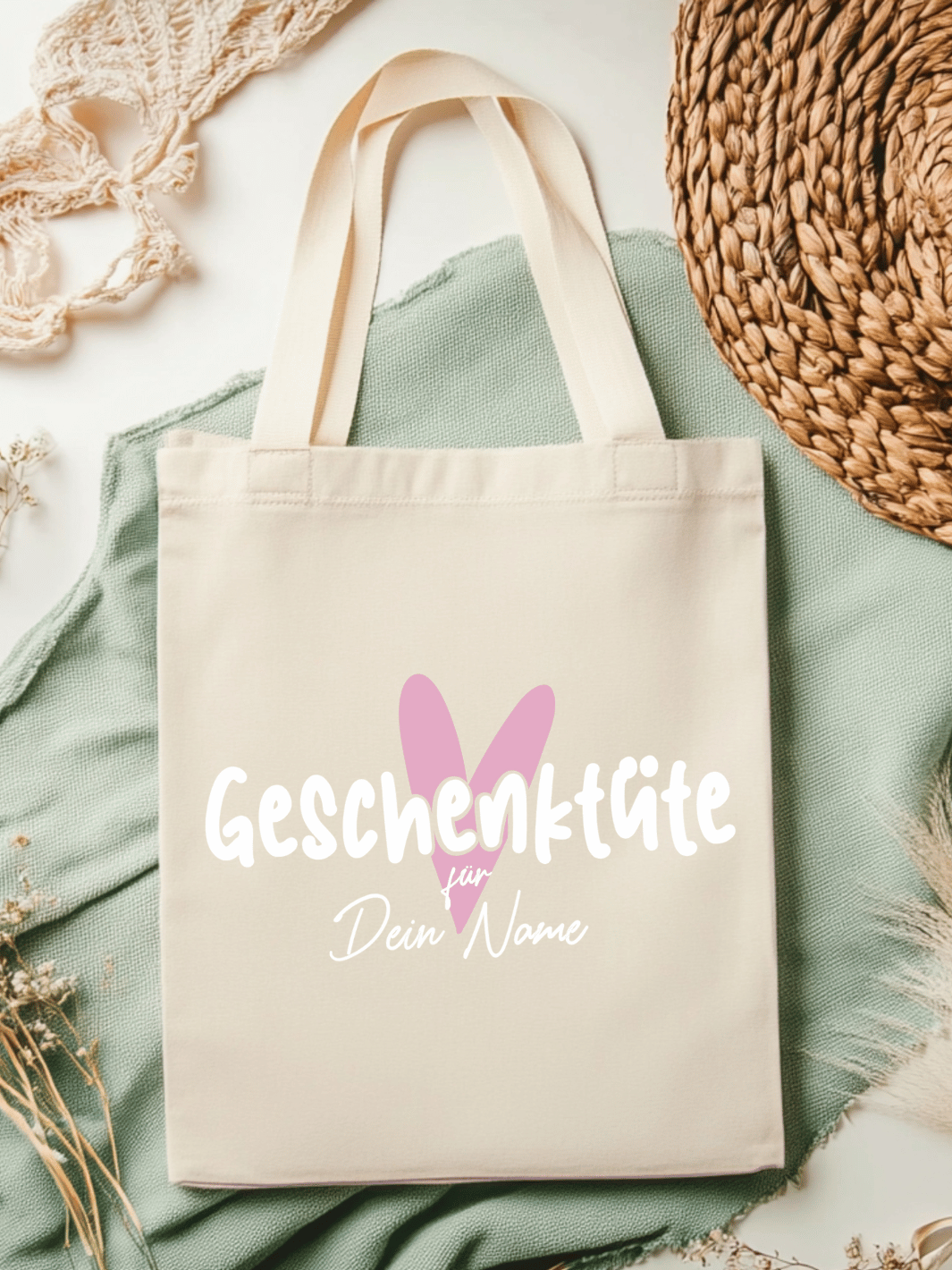 Eine naturfarbene Bio-Baumwolltragetasche, bzw. ein Einkaufsbeutel mit dem Motiv Herz in rosa und Schriftzug Geschenktüte für Dein Name (personalisierbar) , liegt auf einer salbeifarbenen Stoffbahn locker drapiert.