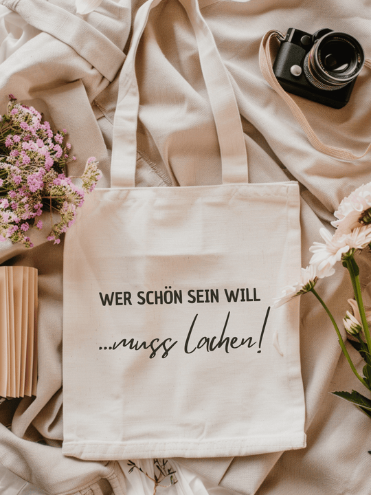 Eine naturfarbene Bio-Baumwolltragetasche, bzw. ein Einkaufsbeutel mit dem Schriftzug: Wer schön sein will muss lachen, liegt auf einer Stoffbahn mit Blumen, Fotokamera und Buch locker drapiert.