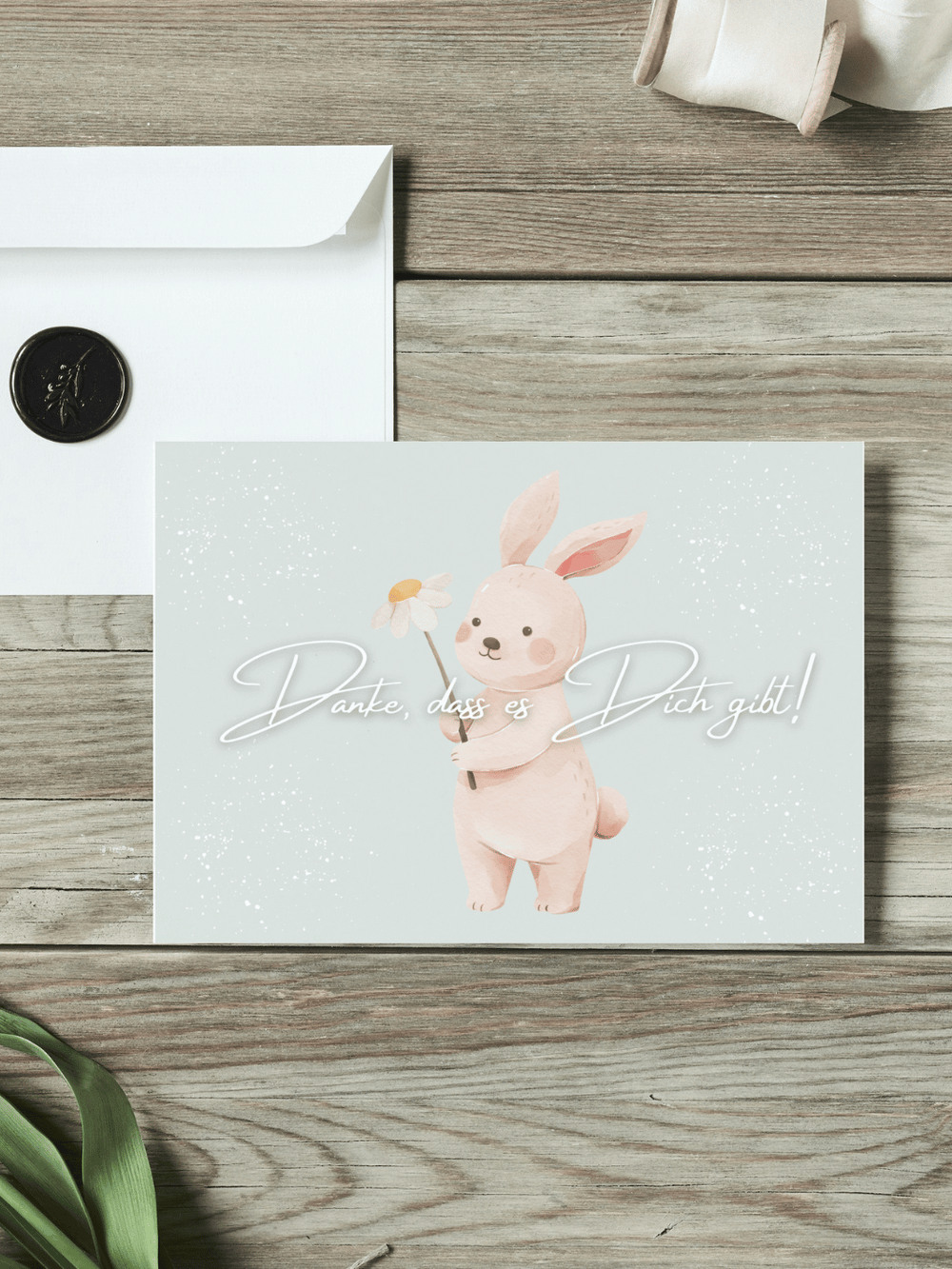 Eine Postkarte mit dem Schriftzug "Danke, dass es dich gibt" über einem Aquarellmotiv Hase mit Blume liegt auf einem Holztisch. Daneben liegt ein Umschlag mit Wachssiegel, sowie eine alte Schere und Stoffband.