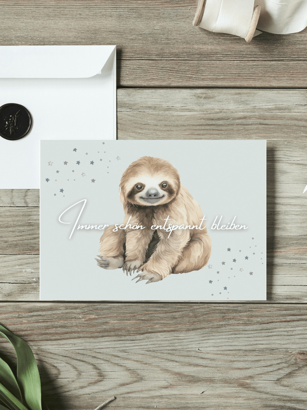 Eine Postkarte mit dem Schriftzug "Immer schön entspannt bleiben" über einem Aquarellmotiv Faultier liegt auf einem Holztisch. Daneben liegt ein Umschlag mit Wachssiegel, sowie eine alte Schere und Stoffband.