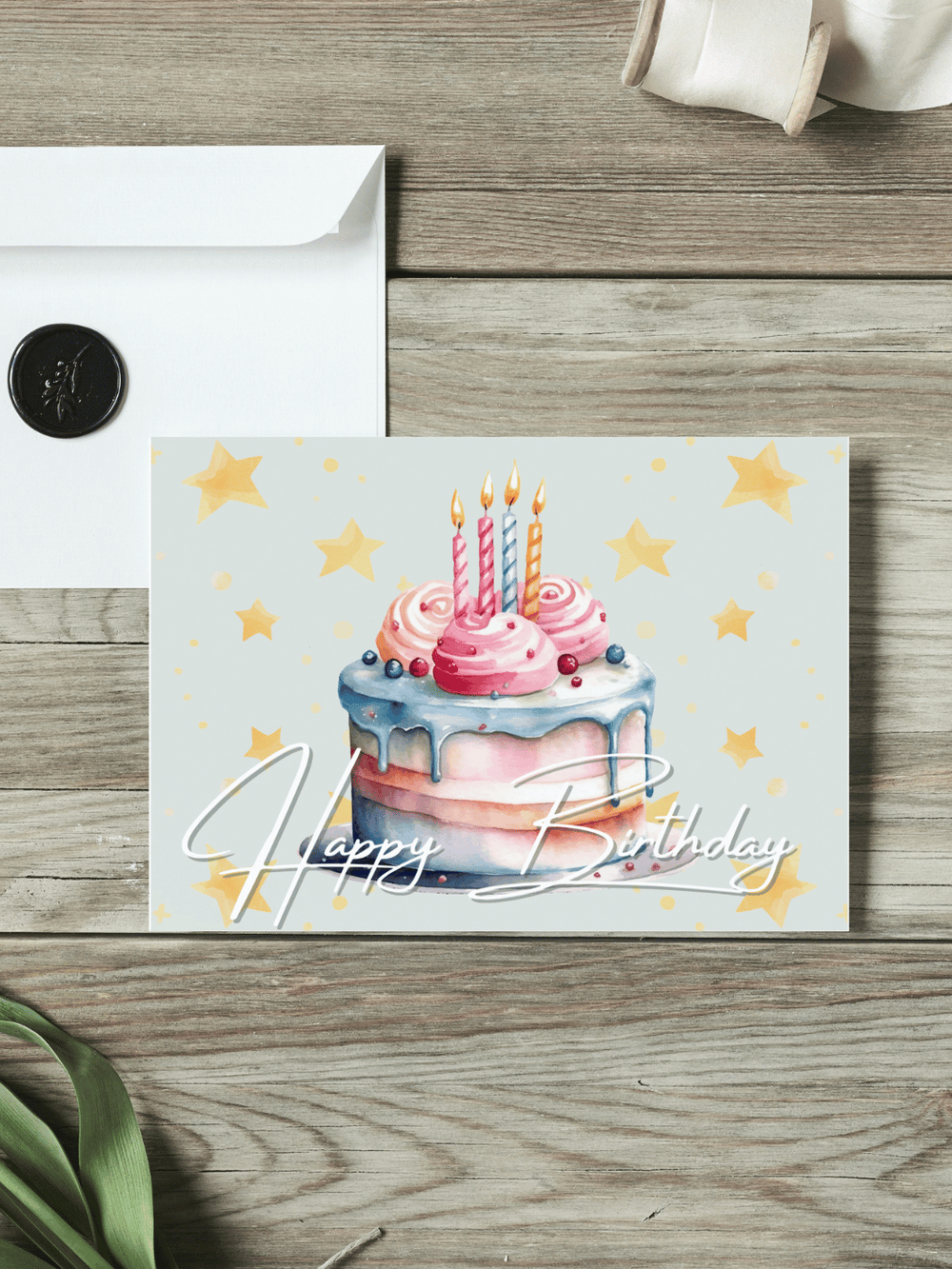 Eine Postkarte mit dem Schriftzug "Happy Birthday" über einem Aquarellmotiv Geburtstagstorte liegt auf einem Holztisch. Daneben liegt ein Umschlag mit Wachssiegel, sowie eine alte Schere und Stoffband.