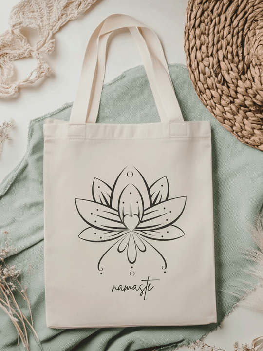 Eine naturfarbene Bio-Baumwolltragetasche, bzw. ein Einkaufsbeutel mit dem Motiv Lotusblume und Schriftzug Namaste, liegt auf einer salbeifarbenen Stoffbahn  locker drapiert.