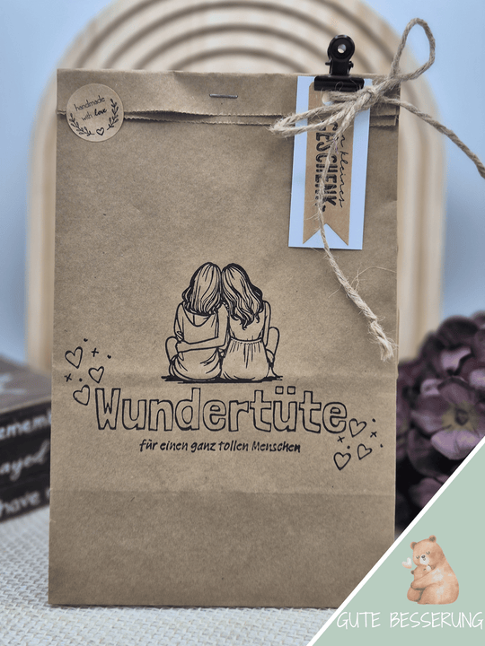 Eine Kraftpapierwundertüte mit einem gestempelten Motiv bestehend aus zwei Frauen, die sich sitzend im Arm halten und der Aufschrift Wundertüte für einen ganz tollen Menschen. Die Wundertüte ist verziert mit einem Handmade-Aufkleber, einer Kordel, Anhängern und einer nostalgischen Büroklammer. Das Thema der Wundertüte ist "Gute Besserung"