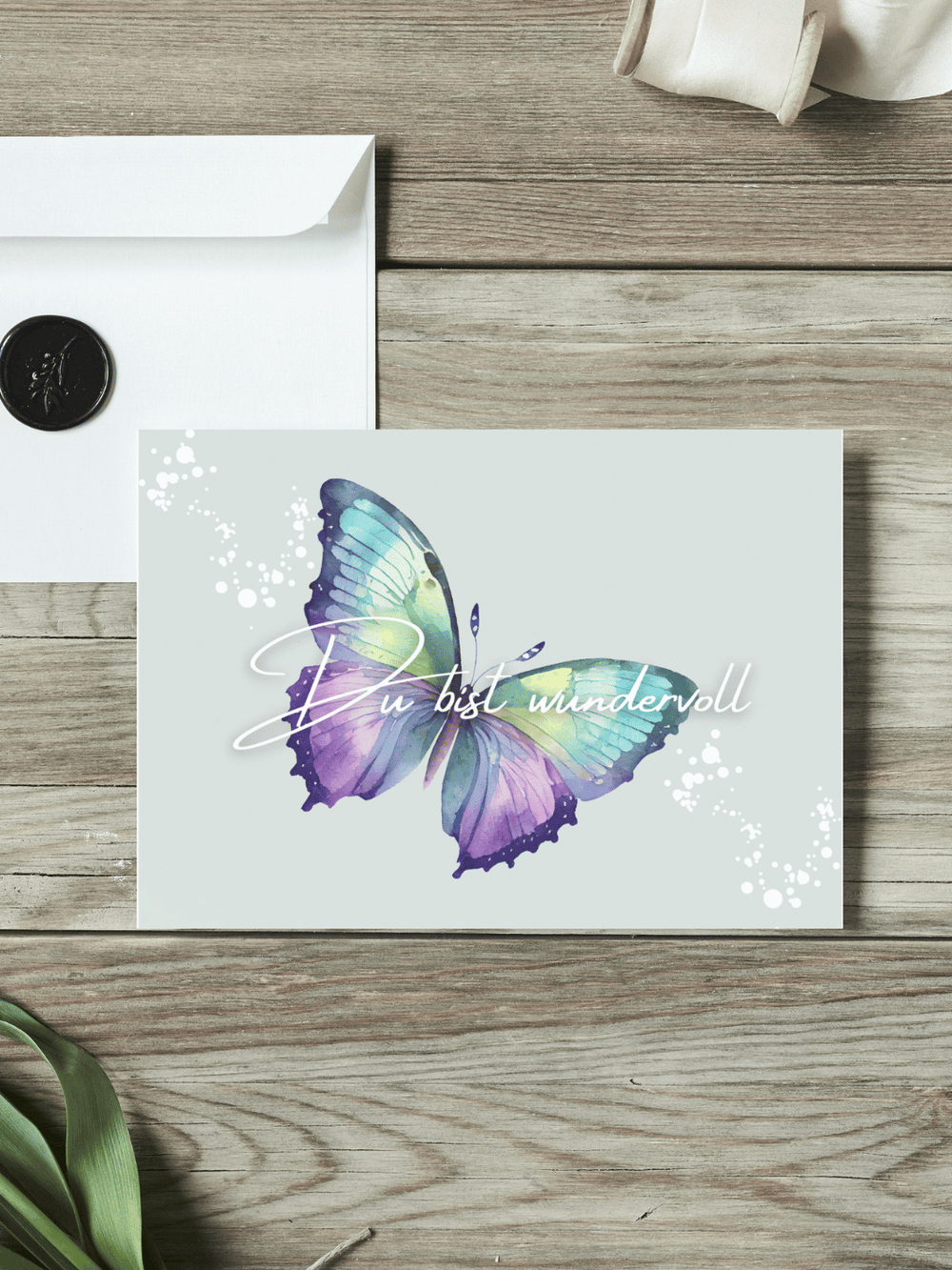 Eine Postkarte mit dem Schriftzug "Du bist wundervoll" über einem Aquarellmotiv bunter Schmetterling liegt auf einem Holztisch. Daneben liegt ein Umschlag mit Wachssiegel, sowie eine alte Schere und Stoffband.
