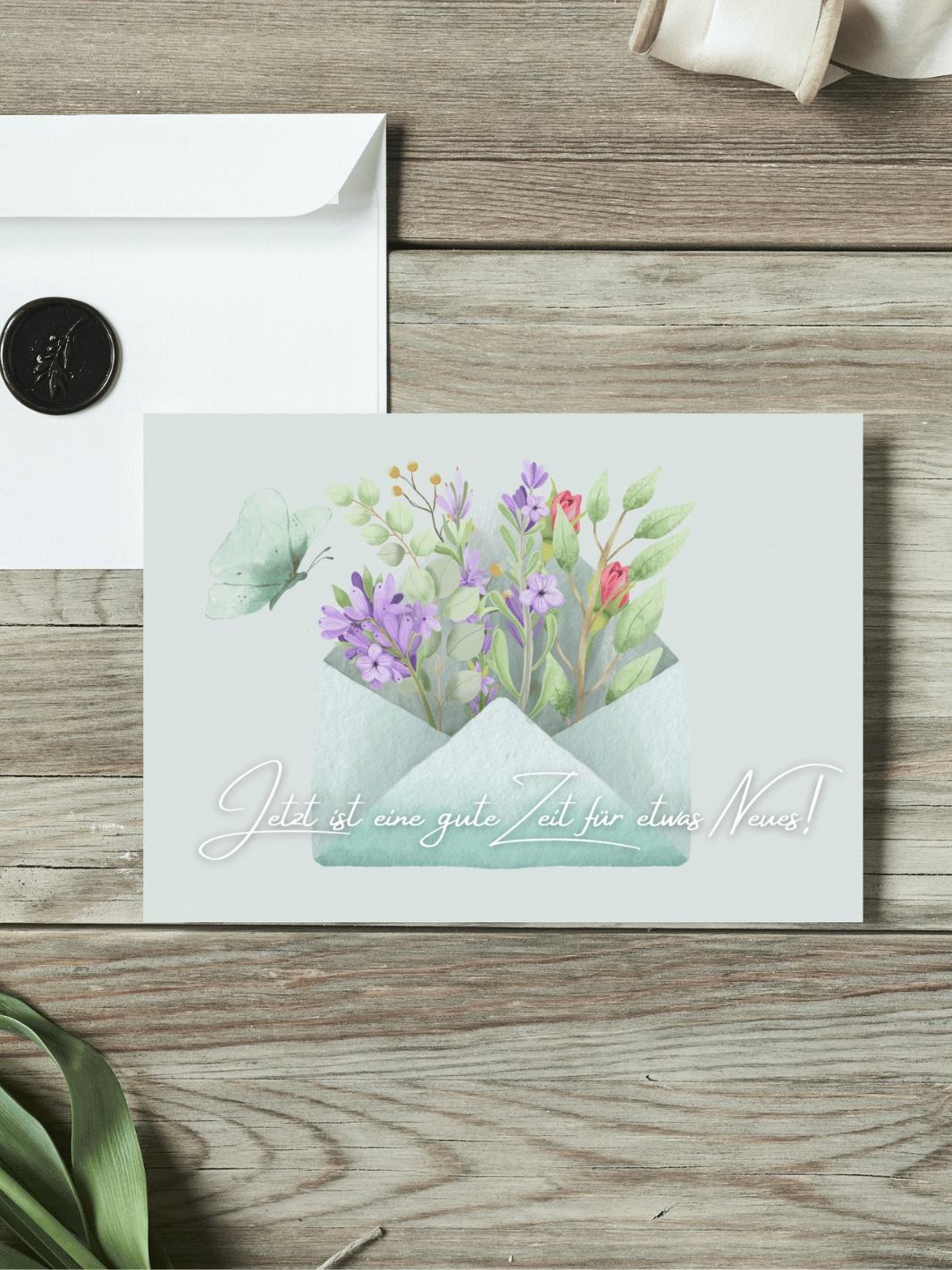 Eine Postkarte mit dem Schriftzug "Jetzt ist eine gute Zeit für etwas Neues" über einem Aquarellmotiv Briefumschlag mit herauswachsenden Blumen und Schmetterling liegt auf einem Holztisch. Daneben liegt ein Umschlag mit Wachssiegel, sowie eine alte Schere und Stoffband.